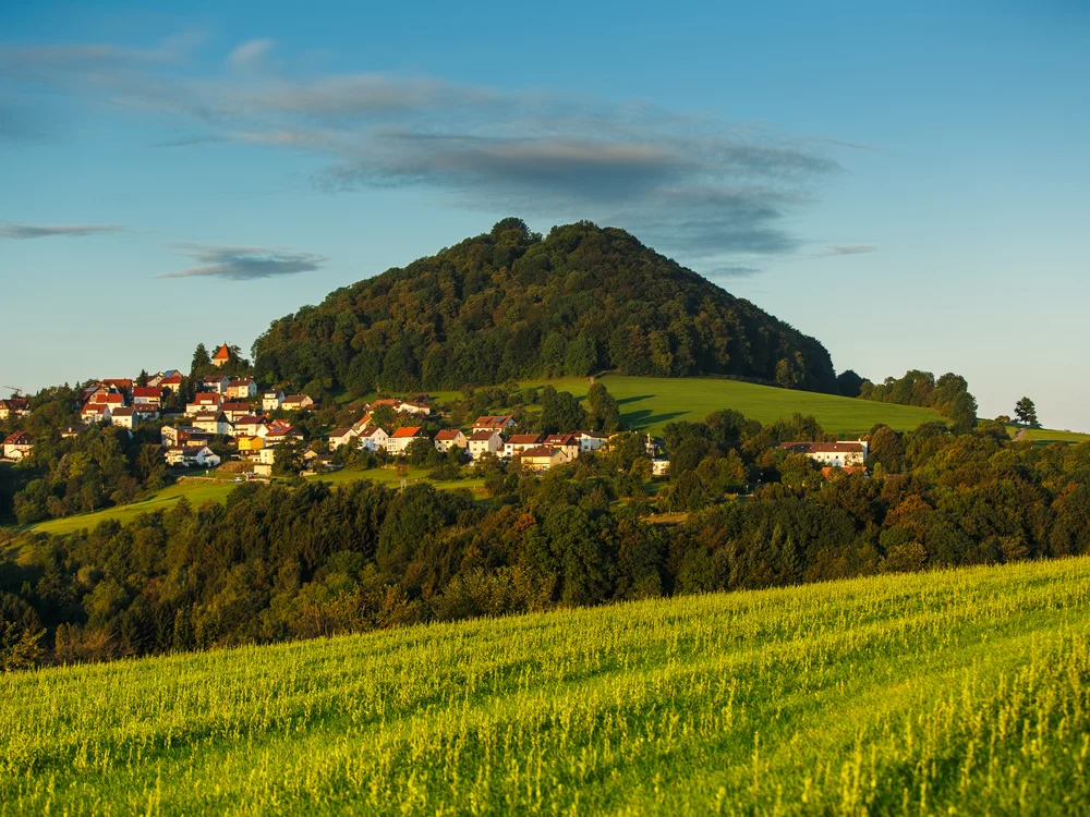 Dorf am Fuß eines bewaldeten Hügels mit grünen Feldern im Vordergrund