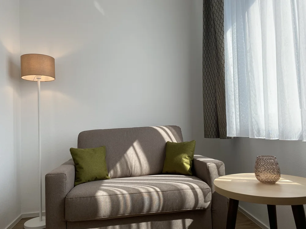 Sessel mit grünen Kissen neben Stehlampe und Tisch am Fenster mit Vorhängen