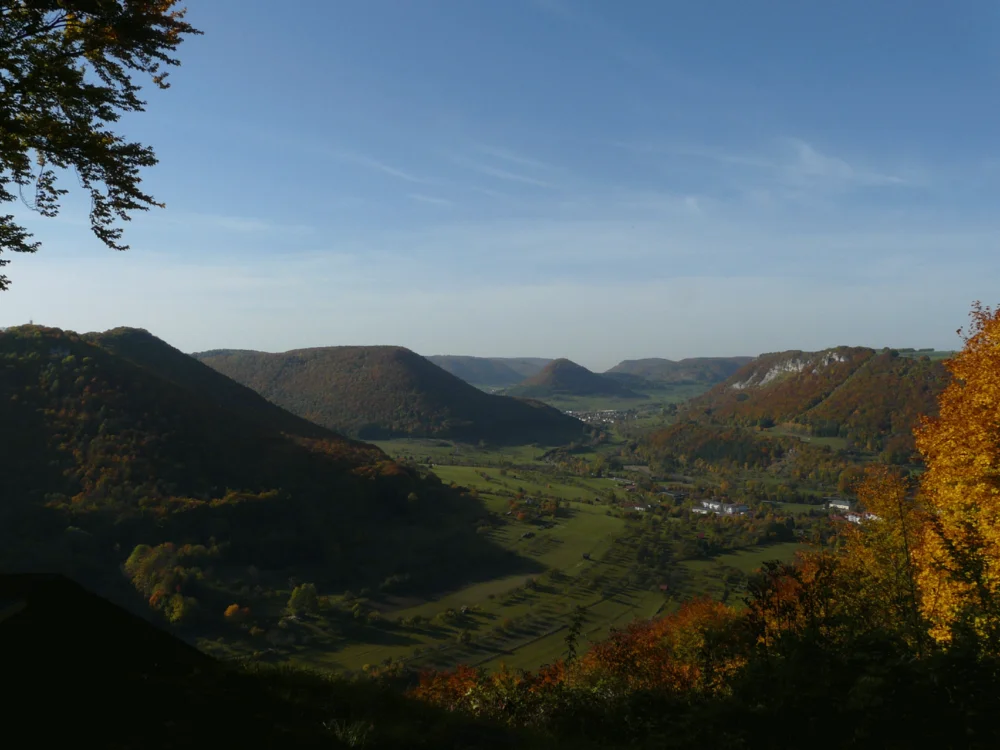 Blick auf ein grünes Tal mit bewaldeten Hügeln unter blauem Himmel